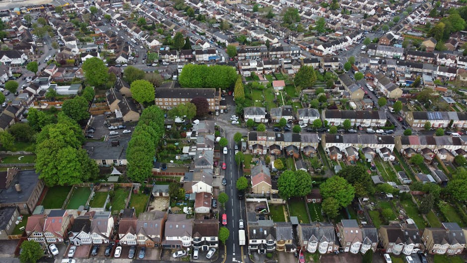 An aerial view of Bramley Road in Cockfosters showing a residential street with houses on both sides and a central lane for vehicles, lined with parked cars and greenery, including trees and gardens. To the left, there is a large green park area with dense trees and open grassy patches. Vehicles are seen moving along the road, while a Man with Van Cockfosters crew is visible loading furniture and packing materials into a van parked near the kerbside, with items such as cardboard boxes, packed furniture, and protective blankets. The image captures the logistical process of home relocation, with a focus on furniture transport and the careful packing of items, reflecting professional removals service within a suburban environment.