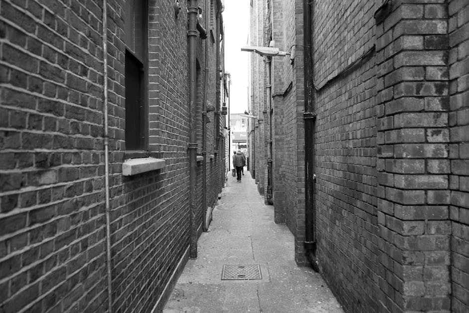A narrow urban alleyway with brick walls on both sides, showing a paved walkway with a square metal drain cover in the center. In the distance, a person dressed in dark clothing is walking away, carry