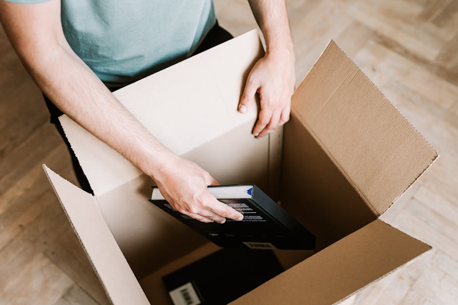 A view inside a cardboard removal box containing a black and white hardcover photo album titled 'HOMEBOOK,' which features images of decorated living rooms, sofas, and interior design details. The box