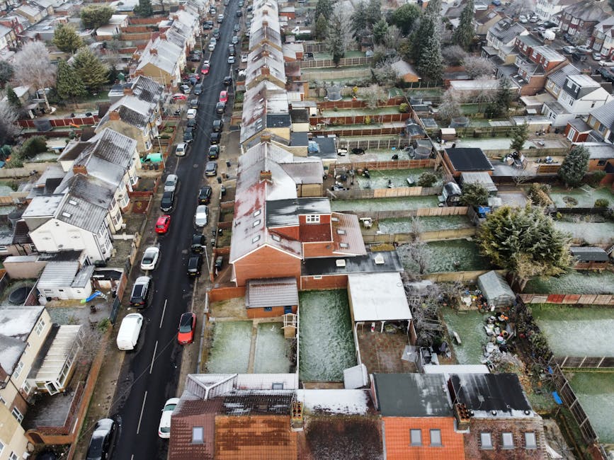 An aerial view of a residential street in Cockfosters showing rows of terraced houses with pitched roofs, some of which have a light dusting of snow. The street is lined with parked cars on both sides, including small to medium-sized vehicles in various colours, and a narrow asphalt roadway runs through the center. Behind the houses, there are numerous back gardens enclosed by wooden fences, some of which contain garden furniture, sheds, and trees, with a mix of evergreen and deciduous varieties. The gardens display different landscaping arrangements, with some areas featuring paved or lawn sections, and others with dense vegetation. The entire scene appears during winter with overcast lighting, and the roofs of the houses exhibit patches of snow or ice. This setting reflects a typical suburban environment suitable for home relocation and furniture transport, often involving moving logistics and packing processes handled by companies like Man with Van Cockfosters.