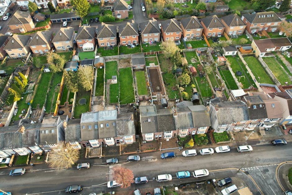 An aerial view of a residential street in Cockfosters, showing a row of terraced houses with individual back gardens. The gardens are bordered by wooden and brick fences, some with trees, sheds, and lawn areas. Several houses have small patios or paved areas at the rear, with visible garden furniture and storage sheds. The street in the foreground has parked cars along each side, with a narrow pavement separating the vehicles from the houses. Bright natural lighting illuminates the scene, highlighting the arrangement of properties and the surrounding greenery. This image captures the typical setting for home relocations and furniture transport operations, as part of a house removal process in line with Enfield Council regulations, with Man with Van Cockfosters providing the moving services.
