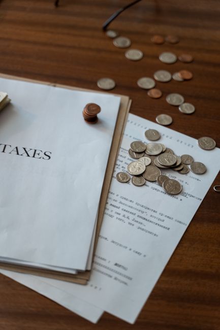 A close-up view of a wooden surface with scattered coins of various denominations, including pennies and smaller silver and copper coins, some piled together and others spread out. Partially visible is a stack of tax-related documents held together with a small copper paperclip, with the word 'TAXES' clearly printed on the top page. The documents are resting on a sheet of paper containing printed text, possibly an invoice or receipt related to home relocation services. The setting suggests an indoor environment, likely a home or office, with natural lighting illuminating the scene. This image reflects the financial aspect of house removals and the importance of clear documentation during furniture transport and moving logistics, as handled by companies like Man with Van Cockfosters.
