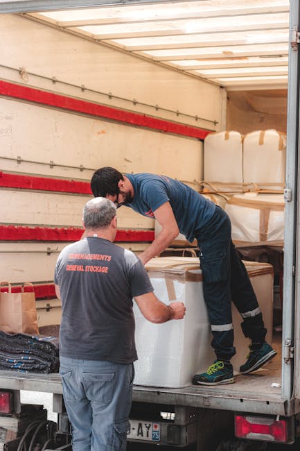 A man wearing blue work overalls and a striped shirt is sitting on the edge of a white van with its door open, inside a property or loading area. He is holding a clipboard and reviewing documents, pos