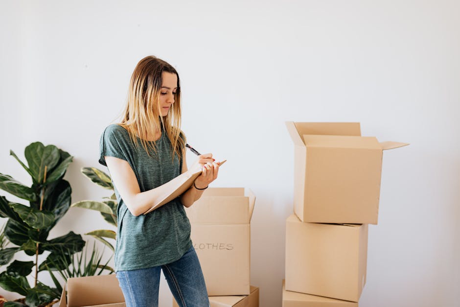 A woman with blonde hair, wearing a dark green T-shirt, is inside a home surrounded by several cardboard boxes, some stacked and open, indicating packing for a home relocation. She is holding a clipboard and writing with a black marker, likely recording inventory or details related to the move. The environment is well-lit, with a white wall in the background and a potted plant visible on the left. The boxes are sealed with brown packing tape, and the scene depicts the packing and moving process, associated with furniture transport and logistics handled by Man with Van Cockfosters as part of their removals services.