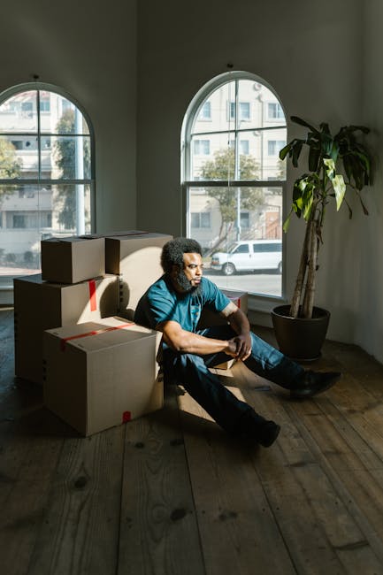 A man with dark curly hair and wearing a blue t-shirt sits on the wooden floor of an interior space during a home relocation process. He is surrounded by several cardboard boxes, some sealed with red 