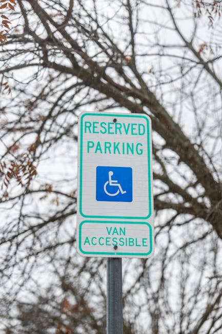 A vertical metal signpost installed on a sidewalk under a tree with bare branches, displaying a reserved parking sign for disabled individuals with a blue wheelchair symbol and the text 'RESERVED PARKING' in green. Below this, a secondary sign indicates 'VAN ACCESSIBLE' in green. The background features an overcast sky and leafless tree branches, with no vehicles or moving equipment visible. The scene is part of an urban area where house removals and moving services, such as those offered by Man with Van Cockfosters, could operate, particularly in contexts involving parking regulations and permits for home relocation logistics.