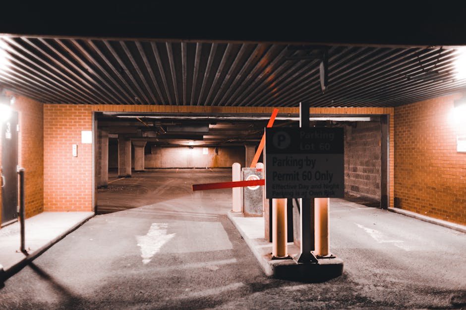 Inside a multi-level underground parking garage with brick walls and a dark, ribbed ceiling. In the foreground, there is a parking barrier arm in a lowered position, made of orange and white plastic, secured by a concrete base. Adjacent to the barrier, a parking sign indicates restrictions for parking permits, with text referencing parking lot number 60 and permit requirements, mounted on a central black pole. The pavement displays a white arrow pointing straight ahead, guiding vehicle movement toward an open, dimly lit section of the garage. To the left, a small, black pedestrian gate is visible near a brick pillar, with subtle lighting illuminating the environment. The scene suggests a secure parking area within a property managed by Man with Van Cockfosters, suitable for loading and unloading furniture during home relocation or moving processes, with visible barriers and signage emphasizing parking control and access guidelines.