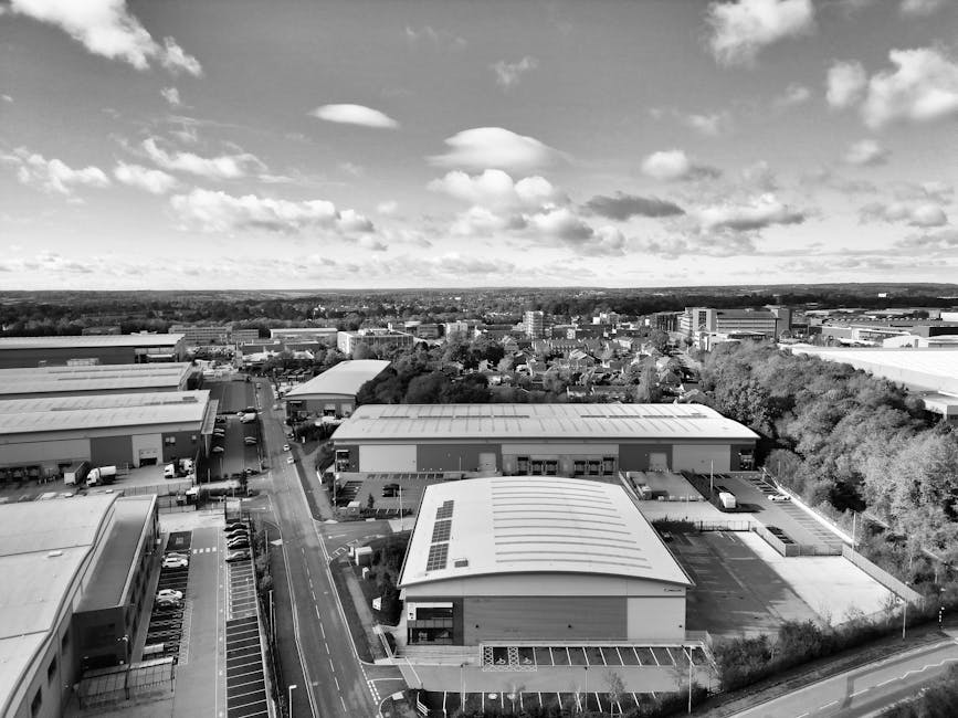 A black and white aerial photograph showing industrial and commercial buildings with flat, metal roofs, some featuring solar panels. The buildings are situated along a paved road with visible parking