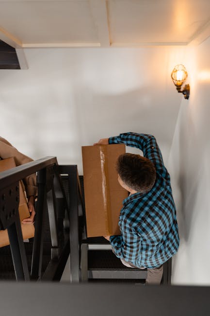 A man wearing a blue and black checkered shirt is standing inside a residential property, carrying a medium-sized cardboard box during a home relocation process. He is positioned near a staircase with black wooden railings and beige cushioned chairs visible on the left side of the image. The room has white walls and ceiling, with a warm yellow wall-mounted light fixture on the right side. The lighting suggests natural daylight combined with the indoor light. The man is holding the box with both hands, preparing to navigate down the staircase, which is part of the furniture transport and packing and moving activities undertaken by Man with Van Cockfosters. The environment appears organized for moving purposes, with minimal visible clutter, and the overall scene depicts a typical moment in the loading process during house removals, emphasizing careful handling of household items in an interior space.