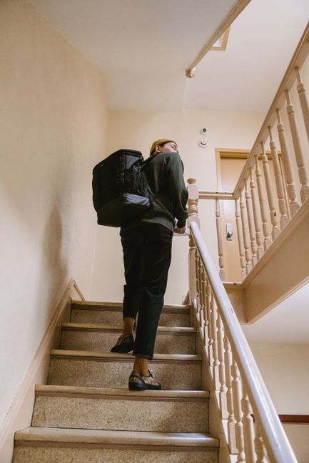 A person wearing dark clothing, including a black jacket and black trousers, is ascending a staircase inside a house. They are carrying a large black backpack and are mid-step on a carpeted stair, with their right foot about to contact a wooden tread. The stairwell features wooden banisters and spindles on the right side, with a cream-coloured wall on the left. The lighting is bright, illuminating the indoor environment. The individual appears to be involved in a home relocation or furniture transport process, possibly part of a moving service such as Man with Van Cockfosters, as seen in the context of staircase challenges faced during house removals in older properties in Cockfosters.