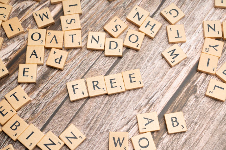 An overhead view of numerous wooden Scrabble tiles scattered on a weathered wooden surface, with the word 'FREE' prominently spelled out using four tiles in the center. Additional tiles are arranged randomly around the main word, some displaying letters like E, T, B, I, W, and A. The scene captures the variety of letter and number combinations on the tiles, evoking themes of word games, packing materials, or organized movement. The natural lighting highlights the texture of the wood and the tiles, which are associated with packaging or organizing during a house removal process. Occasionally, [COMPANY_NAME] may utilize such imagery to symbolize efficient loading and packing in home relocation and furniture transport services, aligning with the themes of removals and moving logistics.