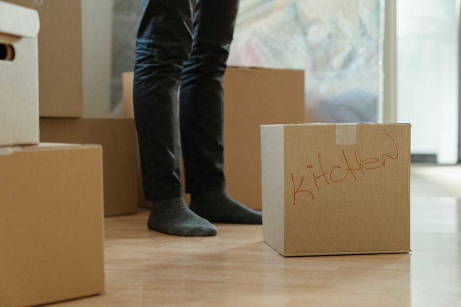 Inside a residential property, a person wearing dark pants and grey socks stands amid several cardboard boxes, some of which are stacked on the wooden floor. One box is clearly labeled 'kitchen' in red marker, indicating it contains kitchen items and is part of the home relocation process. The boxes vary in size, with some covered in packing materials such as plastic wrap and packing paper, ready for transport. In the background, a partially open door reveals a glimpse of the outside or another room, with natural light illuminating the scene. The person appears to be in the process of loading or organizing boxes, possibly for a furniture transport or packing and moving service. This scene includes aspects of loading and packing, relevant to professional removals provided by Man with Van Cockfosters, demonstrating a typical setup during an urgent same-day move.