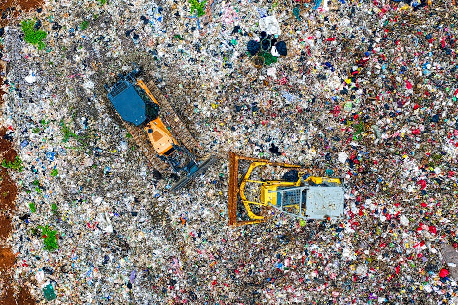 An aerial view of a large landfill site filled with a dense mixture of mixed waste materials including plastic bottles, cardboard boxes, fabric scraps, and various discarded household items. Two heavy construction vehicles are visible; one is a yellow bulldozer spreading and compacting waste, while the other is an orange tracked excavator with a bucket attachment, positioned among the debris. The scene is outdoors, with natural daylight illuminating the accumulated rubbish, which covers the ground extensively and extends to the horizon. The environment appears as a typical waste disposal area, with no immediate structures, and the waste is layered haphazardly. The image reflects the scale of waste generated from home relocations and packing activities, highlighting the importance of professional removals services such as those offered by Man with Van Cockfosters to manage large quantities of bulky waste efficiently during house moves.