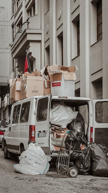 A white commercial van parked on a city street with its rear doors open, loaded with various bulky waste items including large cardboard boxes, some flattened and others still assembled, and black plastic bags filled with rubbish. Cardboard boxes are stacked on top of and around each other in the van's cargo area, with some partially hanging out. The environment shows a multi-storey building façade with windows and architectural details in the background, indicating an urban setting suitable for house removals and furniture transport. A small trolley with additional bags and packaging materials is positioned on the pavement next to the van, suggesting a recent loading process during a home relocation or moving service. The scene captures the typical logistics involved in clearing bulky waste after a move, as handled by professionals like Man with Van Cockfosters.