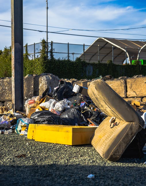 An aerial view of a large landfill site filled with a dense mixture of mixed waste materials including plastic bottles, cardboard boxes, fabric scraps, and various discarded household items. Two heavy construction vehicles are visible; one is a yellow bulldozer spreading and compacting waste, while the other is an orange tracked excavator with a bucket attachment, positioned among the debris. The scene is outdoors, with natural daylight illuminating the accumulated rubbish, which covers the ground extensively and extends to the horizon. The environment appears as a typical waste disposal area, with no immediate structures, and the waste is layered haphazardly. The image reflects the scale of waste generated from home relocations and packing activities, highlighting the importance of professional removals services such as those offered by Man with Van Cockfosters to manage large quantities of bulky waste efficiently during house moves.